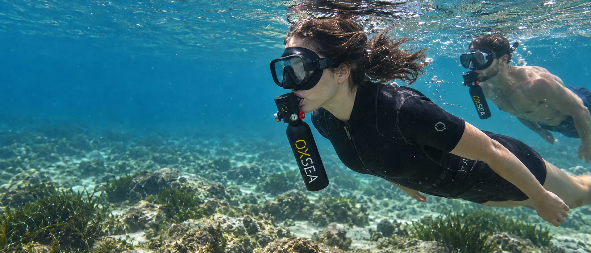 Two divers swim hands-free with their OX500 mouthpieces in place, above a rocky seabed and seagrass