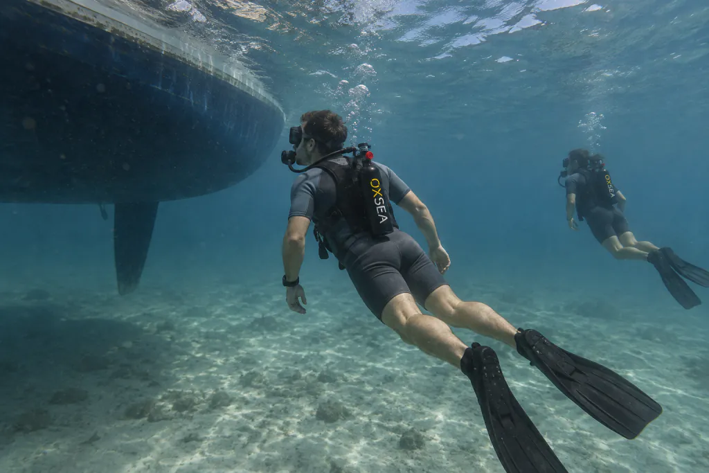 A diver swims under the hull of an anchored sailboat with an OX1000 worn on the back.