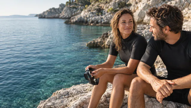 Two friends in wetsuits sitting on the rocks by the sea.